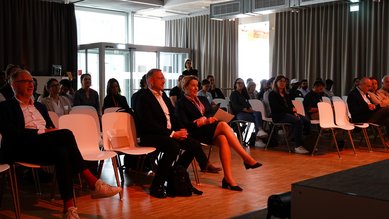 Eine Gruppe von Menschen sitzt in einem Konferenzraum auf weißen Stühlen. Einige Teilnehmer hören aufmerksam zu, während andere auf die Bühne blicken. Der Raum ist gut beleuchtet und hat große Fenster.
