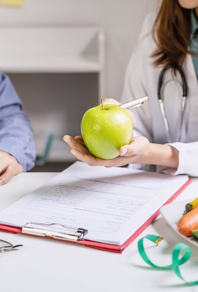 Eine Ärztin hält einen grünen Apfel in der Hand, während sie einem Patienten am Tisch mit gesunden Lebensmitteln und einem Dokument gegenüber sitzt.