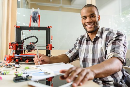 Ein Mann sitzt an einem Tisch mit einem 3D-Drucker, Notizbuch und Tablet, während er einen Stift hält und lächelt.
