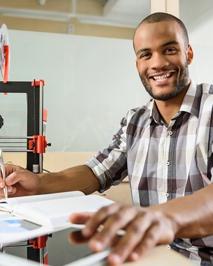 Ein Mann sitzt an einem Tisch mit einem 3D-Drucker, Notizbuch und Tablet, während er einen Stift hält und lächelt.