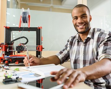 Ein Mann sitzt an einem Tisch mit einem 3D-Drucker, Notizbuch und Tablet, während er einen Stift hält und lächelt.