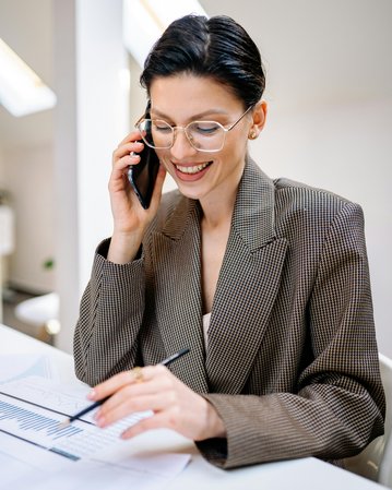 Eine lächelnde Frau mit Brille spricht am Telefon, während sie Notizen auf einem Dokument macht. Sie trägt einen karierten Blazer und sitzt an einem Tisch in einem hellen Raum.