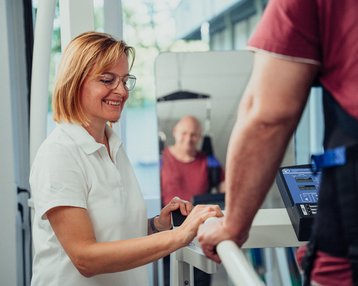Eine lächelnde Frau in einem weißen Polohemd steht an einem Fitnessgerät und spricht mit einem Mann, der an dem Gerät trainiert. Im Hintergrund ist ein weiterer Mann sichtbar.