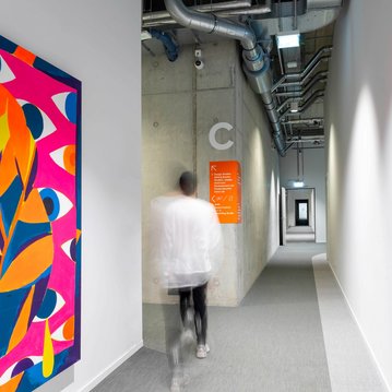 Colorful mural with eye motifs on a bright hallway wall, as a person in a white top walks down the corridor.