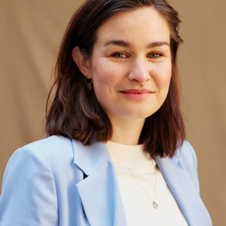 Eine Person mit kurzen, braunen Haaren trägt einen hellblauen Blazer und ein weißes Oberteil. Sie lächelt leicht vor einem neutralen Hintergrund.