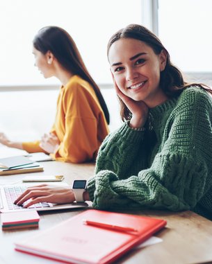 Eine junge Frau mit langen, braunen Haaren sitzt an einem Tisch und lächelt, während sie an einem Laptop arbeitet. Im Hintergrund sind zwei weitere Personen, die ebenfalls an Laptops arbeiten. Rote Notizblätter liegen auf dem Tisch.