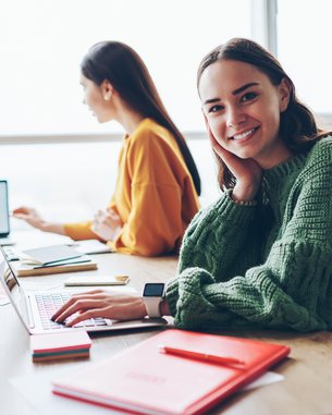 A young woman with long brown hair sits at a table, smiling as she works on a laptop. In the background, two other people are also working on laptops. Red notepads are spread out on the table.
