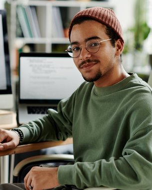 A young man with glasses and a cap sits at a desk, surrounded by computers and office supplies.