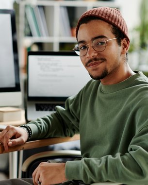 A young man with glasses and a cap sits at a desk, surrounded by computers and office supplies.