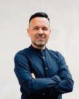 Mann mit kurzem, dunklem Haar und Bart, trägt ein dunkelblaues Hemd und steht mit verschränkten Armen vor einer hellen Wand.