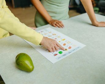 Zwei Personen stehen an einem Tisch und betrachten ein Blatt mit verschiedenen Obst- und Gemüsesorten. Eine Person zeigt mit dem Finger auf ein grünes Gemüse, während eine Avocado neben dem Blatt liegt.