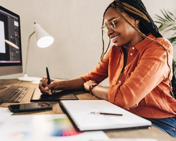 A person with braided hair is working on a graphics tablet while sitting at a computer. On the table, there are notepads and a smartphone. The person is wearing an orange shirt and smiling.