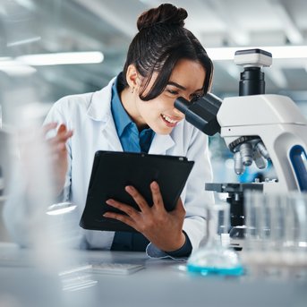 A scientist in a lab is looking through a microscope and recording her observations on a tablet.