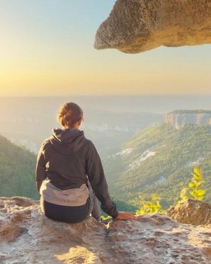 Eine Person sitzt auf einem Felsen in einer Höhle und blickt auf eine Landschaft mit Bergen und einem Sonnenuntergang. Die Szenerie zeigt eine natürliche Öffnung in der Felswand, die den Blick auf die Umgebung freigibt.