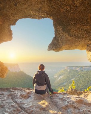 Eine Person sitzt auf einem Felsen in einer Höhle und blickt auf eine Landschaft mit Bergen und einem Sonnenuntergang. Die Szenerie zeigt eine natürliche Öffnung in der Felswand, die den Blick auf die Umgebung freigibt.