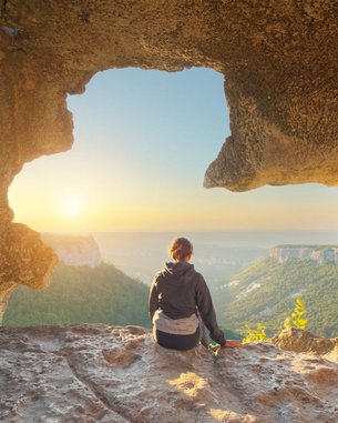 Eine Person sitzt auf einem Felsen in einer Höhle und blickt auf eine Landschaft mit Bergen und einem Sonnenuntergang. Die Szenerie zeigt eine natürliche Öffnung in der Felswand, die den Blick auf die Umgebung freigibt.