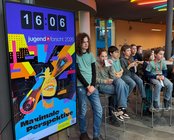 Eine Gruppe von Kindern sitzt auf Barhockern, während ein großes Plakat mit dem Text "Maximale Perspektive" und einer Uhr im Hintergrund zu sehen ist.