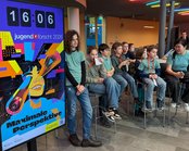 A group of children is sitting on bar stools, while a large poster with the text 'Maximum Perspective' and a clock in the background is visible.