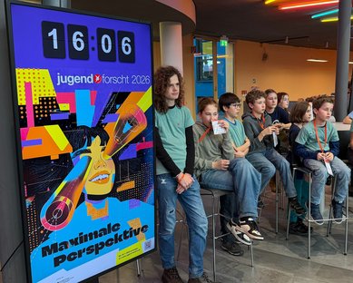 Eine Gruppe von Kindern sitzt auf Barhockern, während ein großes Plakat mit dem Text "Maximale Perspektive" und einer Uhr im Hintergrund zu sehen ist.