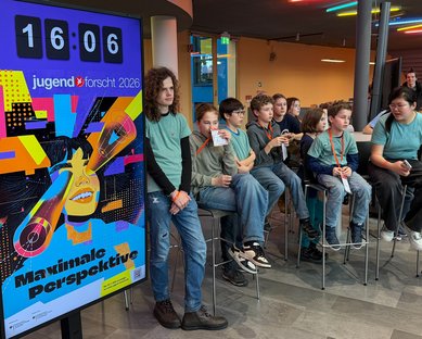 A group of children is sitting on bar stools, while a large poster with the text 'Maximum Perspective' and a clock in the background is visible.