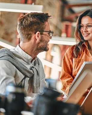 Eine Frau in einer orangefarbenen Bluse hält eine Mappe und spricht mit einem Mann, der eine Brille trägt. Im Hintergrund sind weitere Personen und Büroelemente sichtbar. Die Szene spielt in einem modernen Arbeitsumfeld.