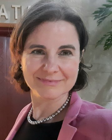 Eine Frau mit kurzen, braunen Haaren und einem rosa Blazer lächelt in die Kamera, während sie eine Perlenkette trägt.