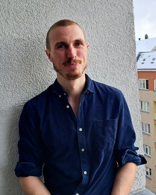 Ein Mann mit kurzen Haaren und Bart steht an einer Wand, trägt ein blaues Hemd und blickt in die Kamera. Im Hintergrund sind Wohngebäude sichtbar.