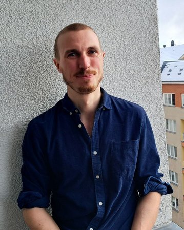 Ein Mann mit kurzen Haaren und Bart steht an einer Wand, trägt ein blaues Hemd und blickt in die Kamera. Im Hintergrund sind Wohngebäude sichtbar.