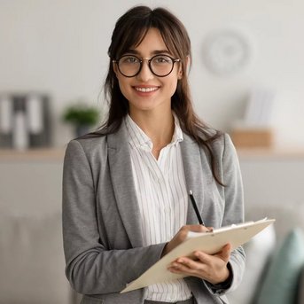 Eine Frau mit Brille in einem grauen Blazer hält ein Notizbuch und lächelt. Im Hintergrund sind Möbel und Pflanzen sichtbar.