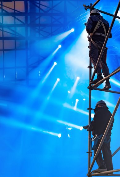 Workers on a scaffold in a brightly lit event arena, surrounded by spotlights and colorful lights.