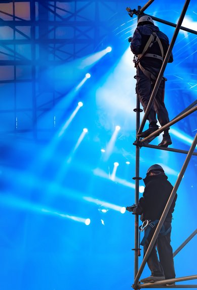 Arbeiter auf einem Gerüst in einer beleuchteten Veranstaltungsarena, umgeben von Scheinwerfern und bunten Lichtern.