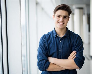 A young man with brown hair stands smiling with his arms crossed in front of a window in a modern building.