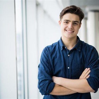 A young man with brown hair stands smiling with his arms crossed in front of a window in a modern building.