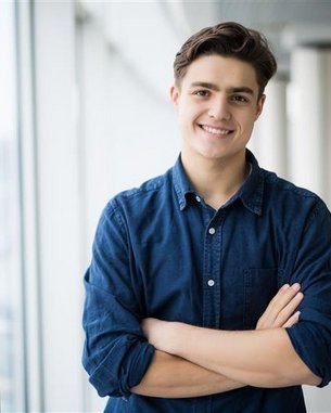 Ein junger Mann mit braunen Haaren steht lächelnd mit verschränkten Armen vor einem Fenster in einem modernen Gebäude.