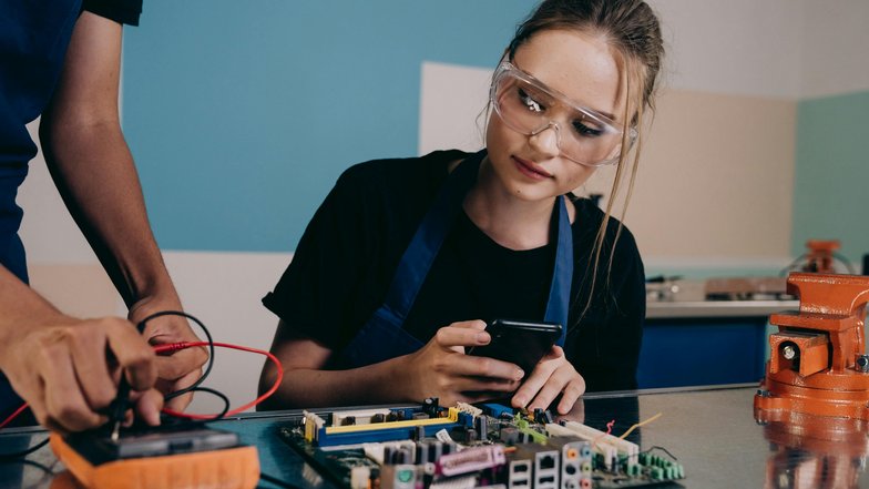 Eine junge Frau mit Schutzbrille betrachtet ein Smartphone, während eine andere Person an einer Leiterplatte arbeitet. Beide sind in einem technischen Umfeld tätig.