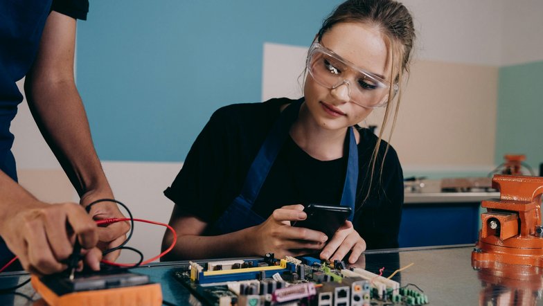 Eine junge Frau mit Schutzbrille betrachtet ein Smartphone, während eine andere Person an einer Leiterplatte arbeitet. Beide sind in einem technischen Umfeld tätig.