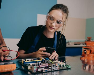 Eine junge Frau mit Schutzbrille betrachtet ein Smartphone, während eine andere Person an einer Leiterplatte arbeitet. Beide sind in einem technischen Umfeld tätig.