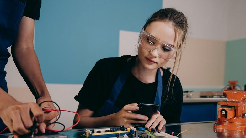 Eine junge Frau mit Schutzbrille betrachtet ein Smartphone, während eine andere Person an einer Leiterplatte arbeitet. Beide sind in einem technischen Umfeld tätig.