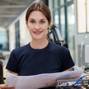 A smiling woman holds blueprints in a modern factory, surrounded by machines and a robotic arm. She is wearing a black T-shirt and has long brown hair.