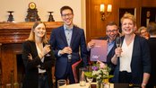 Vier Personen stehen an einem Tisch, halten Gläser und lächeln. Im Hintergrund ist eine elegante Einrichtung mit einem Kamin zu sehen.