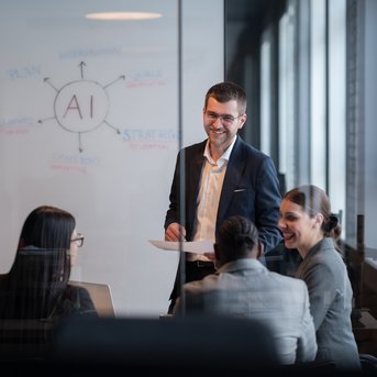 A man is presenting in a conference room in front of a group of four people, while a diagram featuring the word 'AI' is visible in the background.