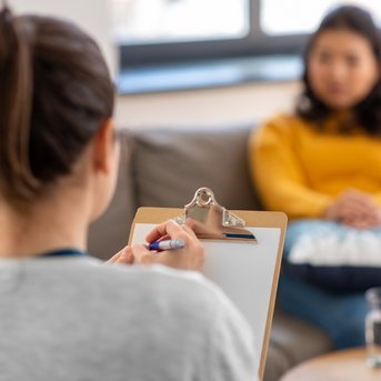 Eine Person sitzt auf einem Sofa und hört einer anderen Person zu, die Notizen auf einem Clipboard macht. Auf dem Tisch steht ein Glas Wasser und eine Schachtel mit Taschentüchern.