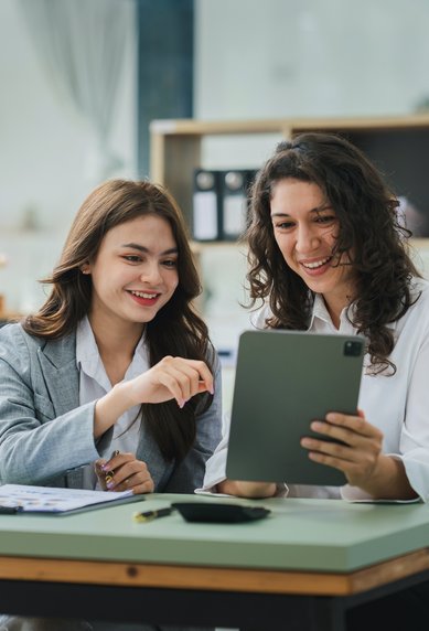 Zwei Frauen sitzen an einem Tisch, schauen auf ein Tablet und lächeln. Ein Laptop und einige Unterlagen liegen auf dem Tisch.