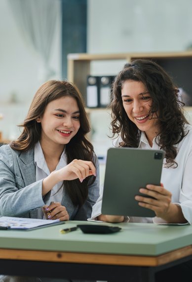 Zwei Frauen sitzen an einem Tisch, schauen auf ein Tablet und lächeln. Ein Laptop und einige Unterlagen liegen auf dem Tisch.