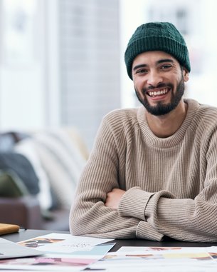Ein lächelnder Mann mit Bart trägt einen grünen Hut und einen beigen Pullover, sitzt an einem Tisch mit einem Laptop und Unterlagen.