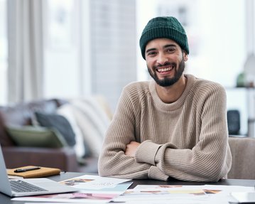 Ein lächelnder Mann mit Bart trägt einen grünen Hut und einen beigen Pullover, sitzt an einem Tisch mit einem Laptop und Unterlagen.