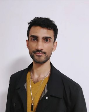 Ein junger Mann mit kurzem, dunklem Haar trägt ein schwarzes Oberteil und eine goldene Kette. Der Hintergrund ist neutral und einfarbig.