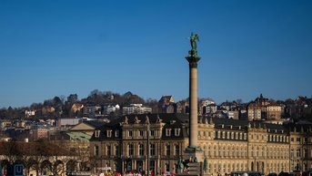 A large crowd gathers in a square, surrounded by historic buildings and a tall monument featuring a statue.