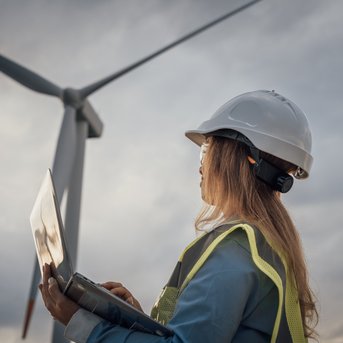 A person in protective gear and a helmet is working with a laptop in front of a wind turbine.