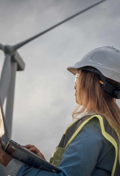 A person in protective gear and a helmet is working with a laptop in front of a wind turbine.
